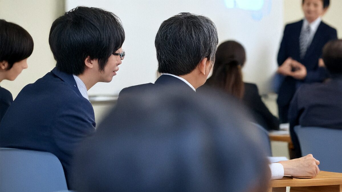 ビジネスセミナーの風景。講師がスクリーンを使って説明し、参加者が熱心にメモを取っている様子。